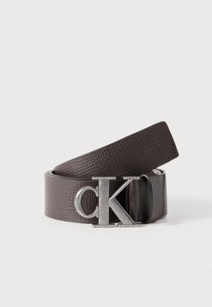 Bruine leren riem met een gestructureerde afwerking, voorzien van een zilverkleurige gespsluiting met het "CK"-logo. De riem heeft een gladde binnenzijde en een plat randontwerp.