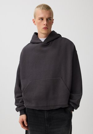 Young man with short blond hair wearing a loose dark gray hoodie and black pants, looking to the side against a plain background.