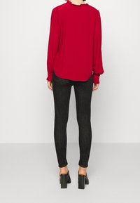 Femme portant un chemisier rouge à manches longues, un jean skinny noir et des chaussures à talons noirs, vue de dos sur un fond blanc.