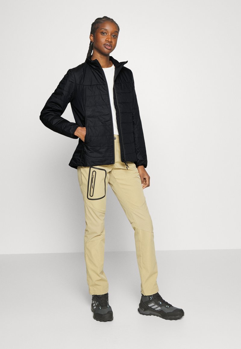 Schwarze Steppjacke mit vertikalem Steppmuster, einem hohen Kragen, kombiniert mit beigen Hosen mit Reißverschlusstasche und schwarzen Sportschuhen.