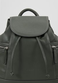 Sac à dos en cuir vert foncé avec un rabat, fermeture à cordon, poches zippées à l'avant et détails de couture blanche. Logo de la marque embossé en bas.