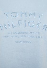 Vaaleansininen puuvillakangas, jossa on kirjailtu teksti: "TOMMY HILFIGER," osoite ja vuosi hillityssä sävyssä. Selkeä ja minimalistinen muotoilu.