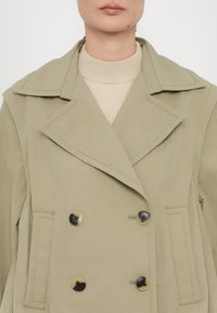 Femme portant un trench-coat beige à double boutonnage avec de grands revers sur un pull à col roulé beige clair.