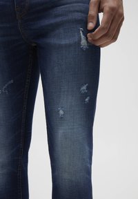 Vaqueros de mezclilla azul oscuro con desgastes deshilachados y decoloración ligera. Vista de cerca que resalta la textura de la tela y el detalle de las costuras a lo largo de las uniones.