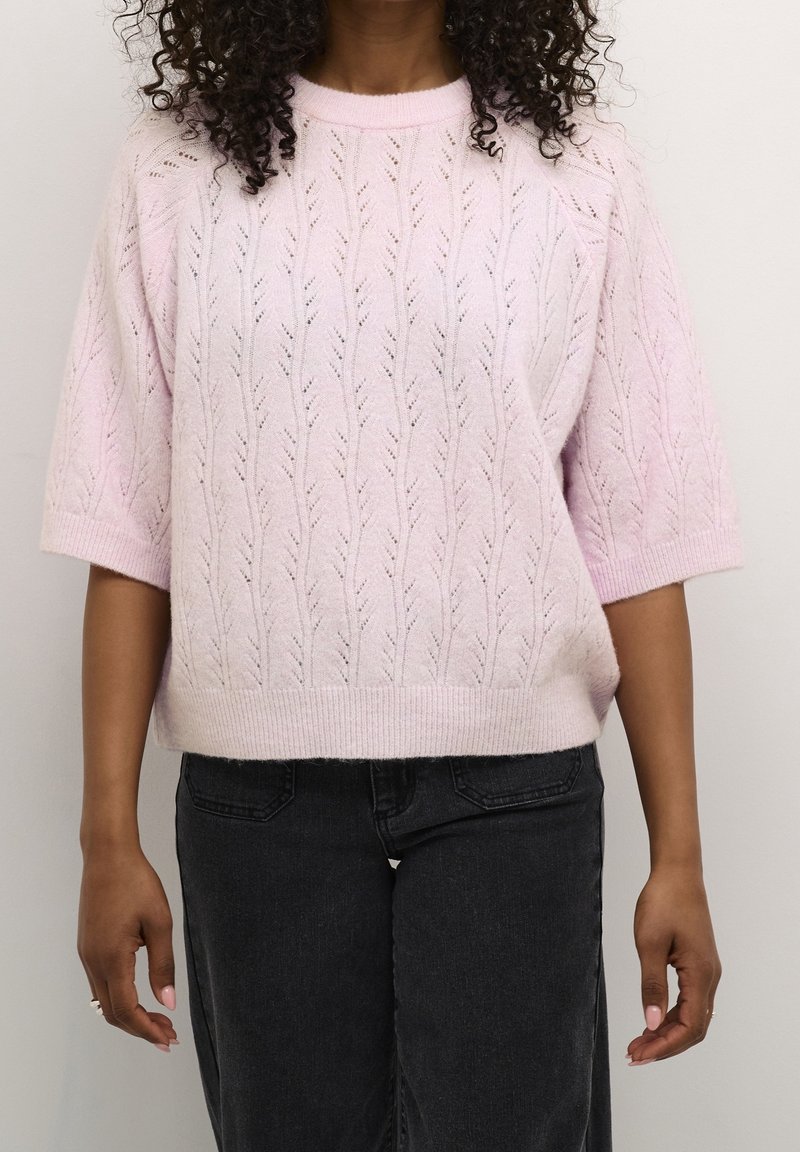 Femme portant un pull en maille texturée rose clair à manches courtes et un jean gris foncé, debout devant un fond uni.