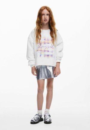 Sweat-shirt blanc avec un motif à carreaux coloré et du texte, associé à une jupe argentée brillante et des chaussures métalliques à brides.