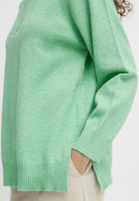 Pull en maille verte avec poignets et ourlet côtelés, doté d'une fente latérale pour faciliter les mouvements et d'une coupe décontractée. Tissu léger et texturé.