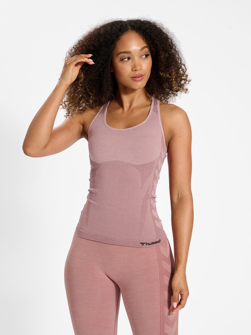 Vrouw met krullend haar draagt bijpassende mauve mouwloze top en legging, staat met één hand haar haar aanrakend en kijkt opzij.
