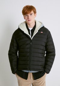 Chaqueta acolchada negra con capucha forrada de Sherpa, cierre de cremallera y diseño acolchado. Llevada sobre una camisa negra de botones y pantalones de pana.