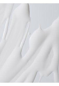 Sustancia cremosa blanca con una textura brillante, extendida de manera irregular sobre una superficie gris clara, con diferentes grosores y bordes suaves.