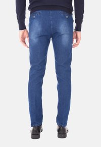 Jeans in denim blu slim-fit con tasche posteriori e un leggero effetto wash. Indossati con scarpe nere, che mostrano una trama di tessuto liscia.