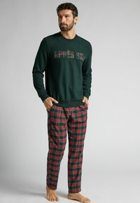 Sweatshirt vert foncé avec "APRES SKI" dans un motif à carreaux. Accompagné d'un pantalon de pyjama à carreaux rouges et verts. Pas de chaussures portées.