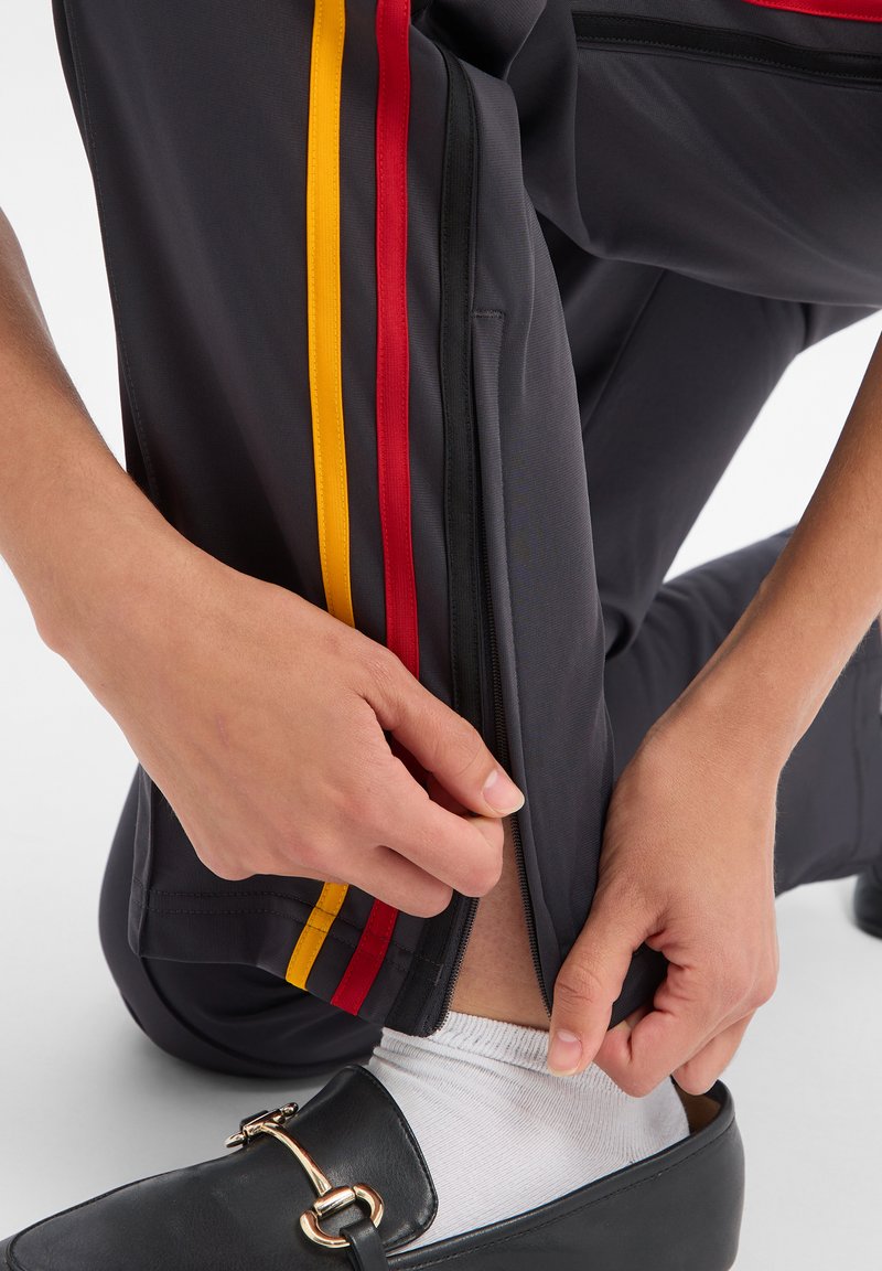 Des mains fermant la fermeture éclair à la cheville d'un pantalon foncé avec des bandes latérales rouges et jaunes, montrant une chaussette blanche et une chaussure mocassin noire.