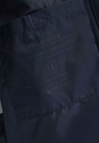 Ciemnoniebieska metka z napisem "POLO CLUB", ikonami pielęgnacji, oznaczającymi prosty, dopasowany krój, właściwości termiczne, wodoodporność oraz poliester pochodzący z recyklingu.