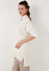 Creme strik uden ærmer med sideslids, en afslappet pasform og ribbede kanter. Båret over en hvid skjorte med oprullede ærmer og neutrale bukser.