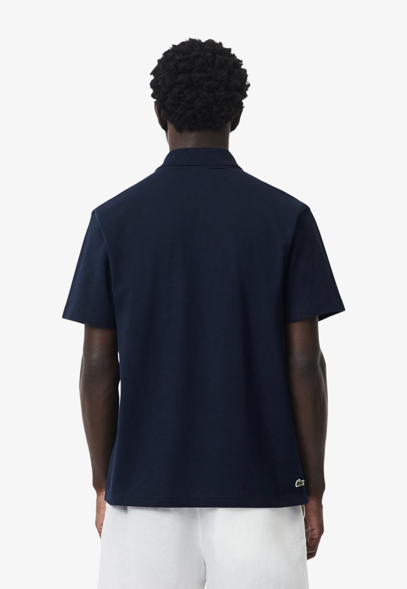 Homme aux cheveux courts et bouclés portant un polo bleu marine uni à manches courtes et un short blanc, vu de dos.