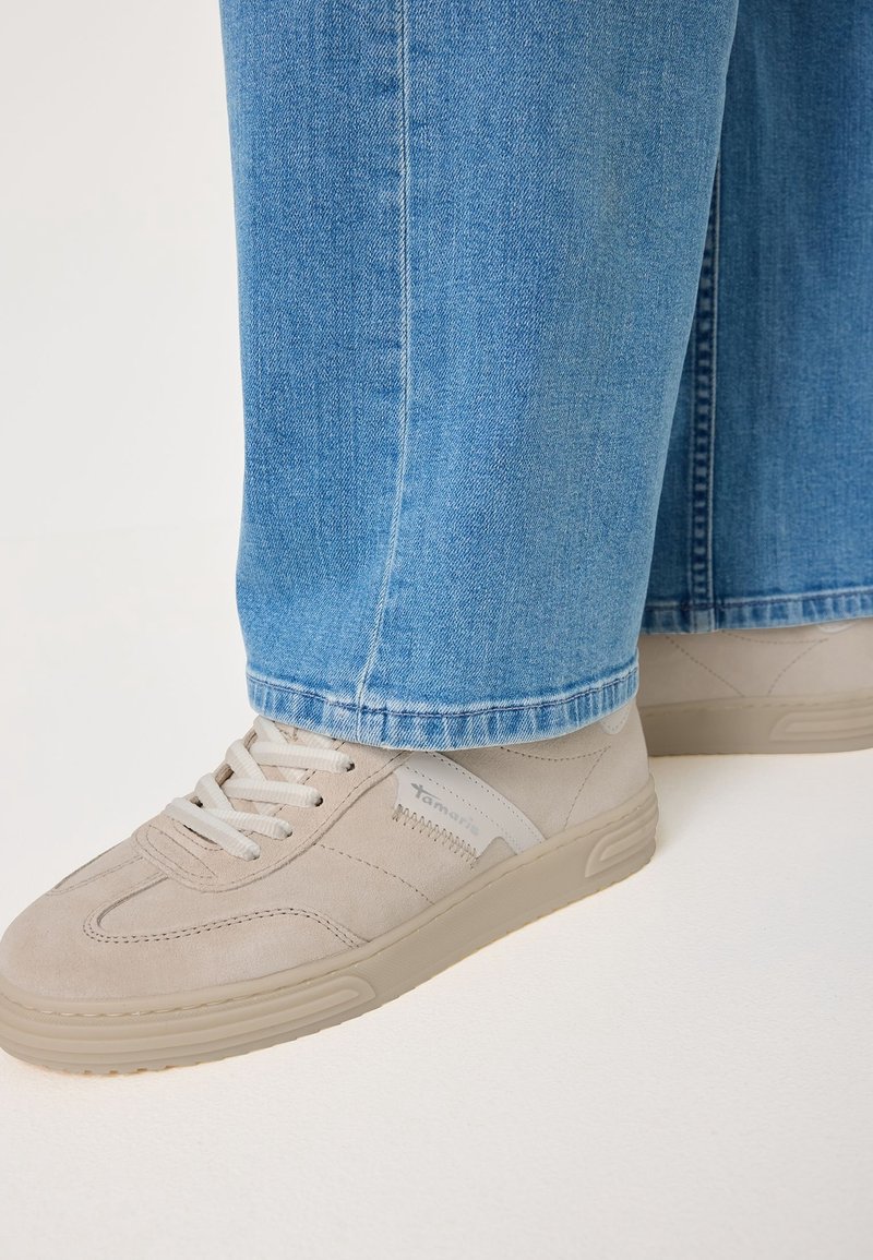 Beżowe zamszowe sneakersy z szarymi akcentami i białymi sznurowadłami, zestawione z jasnoniebieskimi dżinsami o prostym kroju i przeszywanym hemie.