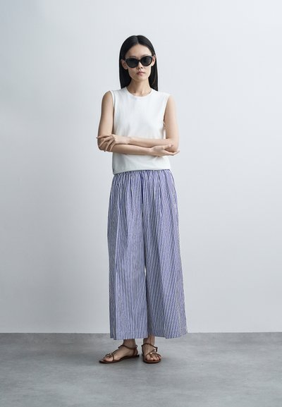 Femme portant des lunettes de soleil noires, un top blanc sans manches, un pantalon large à rayures verticales bleu et blanc, et des sandales marron, debout les bras croisés.