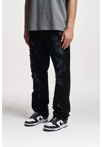 Jean en denim noir déchiré avec une coupe décontractée, présentant de multiples déchirures et des bords effilochés, associés à des baskets noir et blanc.