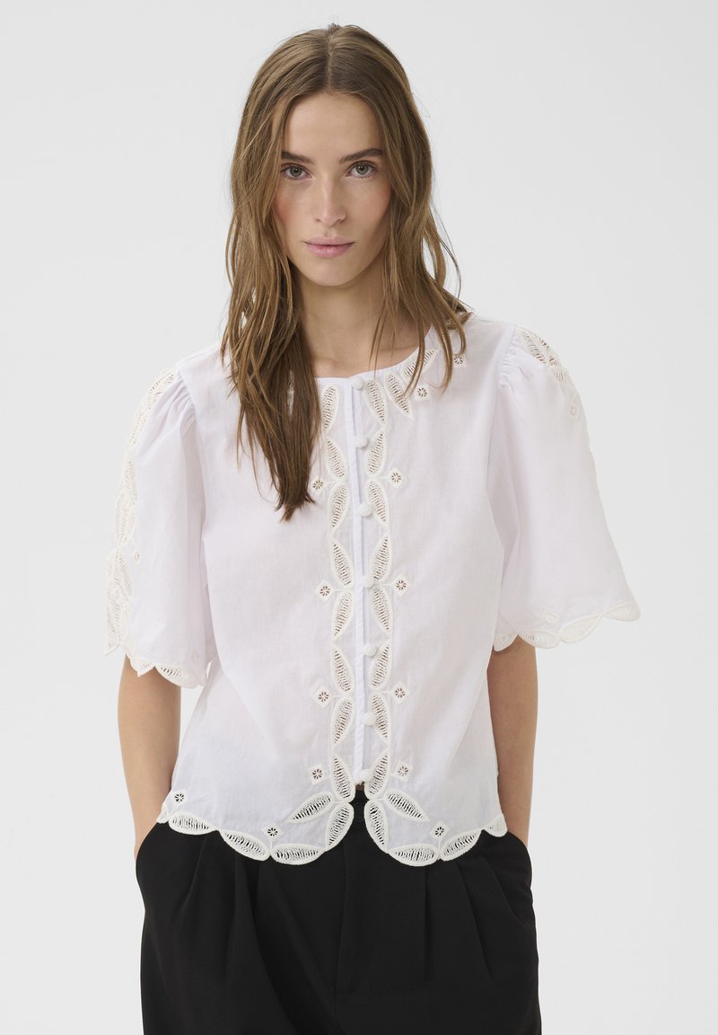 Jeune femme aux longs cheveux bruns portant un chemisier blanc brodé avec des bords en feston et un pantalon noir, les mains dans les poches.