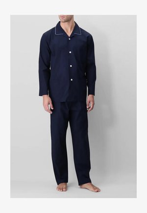 Uomo in piedi a piedi nudi che indossa un pigiama blu navy con maniche lunghe e bottoni e pantaloni da pigiama abbinati e larghi, su uno sfondo semplice.