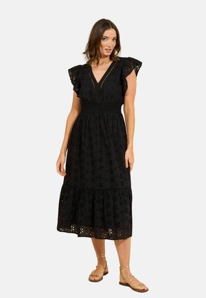 Žena oblečená v čiernych midi šatách s kvetinovým dielikovým vzorom, s priesvitnými rukávmi, výstrihom do V, zdobeným pásom a zlatými sandálmi na plochom opätku so remienkami.