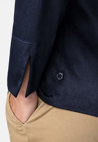 Marineblaues Kleidungsstück mit einem seitlichen Schlitz am Ärmel und einem runden schwarzen Knopf mit erhabenem Text, kombiniert mit helltanfarbener Hose.