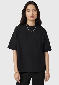 Jonge vrouw met lang donker haar, gekleed in een eenvoudige oversized zwarte t-shirt, zwarte broek, een tweekleurige ketting en armbanden op een lichtgrijze achtergrond.