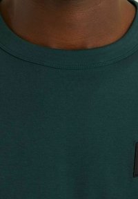 Mörkgrön bomullströja med ribbad rund halsringning och en liten svart etikett på framsidan. Mjuk textur, minimalistisk design.