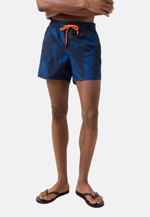 Mand iført marineblå badeshorts med orange snor og sorte klipklappere, stående med armene over kors mod en ensfarvet baggrund.