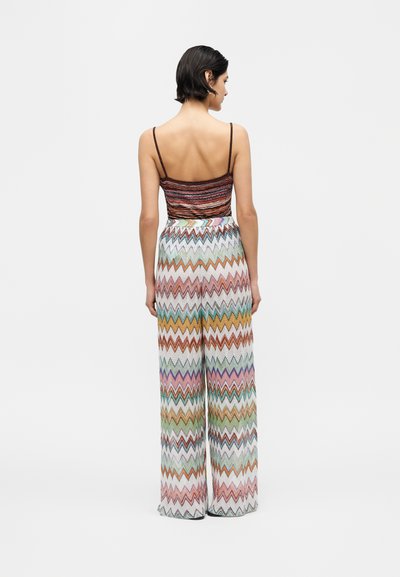 Γυναίκα με κοντά σκουρόχρωμα μαλλιά που φοράει αμάνικο καφέ ριγέ τοπ και ψηλόμεσο παντελόνι με φαρδιά μπατζάκια και πολύχρωμο ζιγκ ζαγκ μοτίβο, με την πλάτη γυρισμένη.