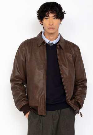 Veste en cuir marron avec un col, texture matelassée, fermeture éclair et poches latérales, portée par-dessus un pull bleu marine et une chemise à motifs.