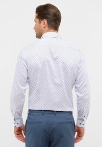 Camicia bianca a maniche lunghe con un motivo geometrico blu, realizzata in morbido cotone, design aderente e polsi con bottoni, abbinata a pantaloni blu scuro.