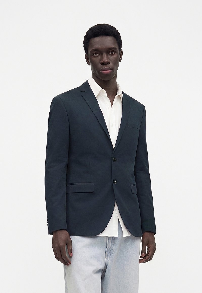 Giovane uomo con blazer scuro, camicia bianca e jeans azzurri chiari, in piedi davanti a uno sfondo chiaro e uniforme.