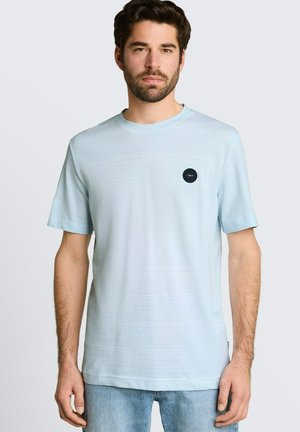 Mann mit dunklen Haaren und Bart, der ein hellblaues T-Shirt mit kurzen Ärmeln mit einem kleinen schwarzen runden Logo auf der Brust und hellblaue Jeans trägt.