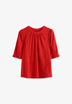 Punainen kuvioitu pusero, jossa on timantinmuotoinen silmukkakuvio, pyöreä pääntie ja kolmen neljänneksen mittaiset liehukehihat, joissa kootut hihansuut.