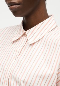 Nahaufnahme einer Person, die ein weißes Hemd mit hellrosa vertikalen Streifen und spitzem Kragen trägt, oben zugeknöpft.