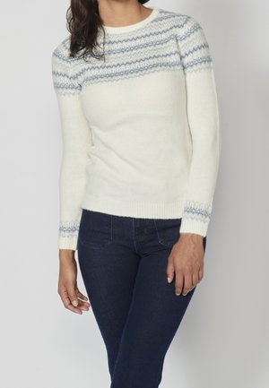 Femme portant un pull blanc avec des rayures bleues à motifs sur la poitrine et les poignets, associé à un jean bleu foncé.