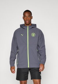 Siva dukserica s kapuljačom i neon zelenim detaljima i zatvaračem, s logotipom Manchester Cityja na prsima, lagani materijal i elastične manžetne.