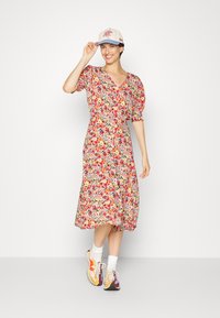 Vestido floral com mangas curtas bufantes, padrão de flores multicoloridas, até ao joelho. Combinado com um chapéu bege e ténis multicoloridos.