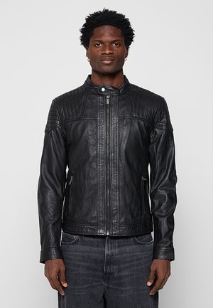 Mann trägt eine schwarze, gesteppte Lederjacke mit Reißverschluss und Druckknopfkragen, kombiniert mit dunkelgrauen Jeans, steht vor einem schlichten Hintergrund.