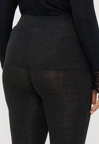 Juodos leggingai su blizgančiu, taškuotu raštu. Aukšta juosmens juosta ir elastinga medžiaga, demonstruojanti artimą prigludimą su tekstūruota paviršiaus.