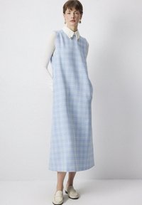 Rochie fără mâneci, cu carouri albastre, dotată cu buzunare laterale, realizată dintr-un material texturat. Prezintă o cămașă albă cu guler dedesubt și loafers crem.