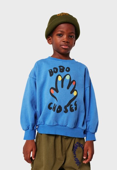 Felpa blu con grafica di mani colorate e testo "BOBO CHOSES", con maniche lunghe e polsini elasticizzati. Indossata con pantaloni verde oliva.