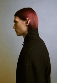 Fekete, magas gallérú dzseki, amelynek formája jól meghatározott, sima textúrával rendelkezik. A modellen egyenes, vállig érő sötétvörös haj van, profilból látható.