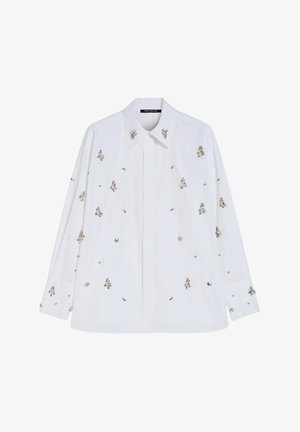 Camicia bianca in cotone con colletto, decorata con ornamenti argentati e piccoli motivi floreali su tutta la superficie. Maniche lunghe con polsini a bottoni.