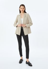 Beige skräddarsydd blazer med två guldknappar, bärs över en vit topp och svarta byxor, ihop med svarta loafers, mot en enhetlig bakgrund.