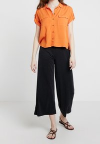 Pomarańczowa koszula na krótki rękaw z guzikami i dwoma kieszeniami na piersi, zestawiona z luźnymi, czarnymi spodniami o szerokich nogawkach oraz brązowymi, płaskimi sandałami.