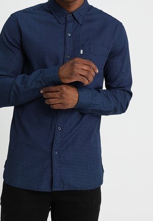 Homme portant une chemise à carreaux bleue à manches longues boutonnée, ajustant une manchette avec l'autre main sur fond blanc.