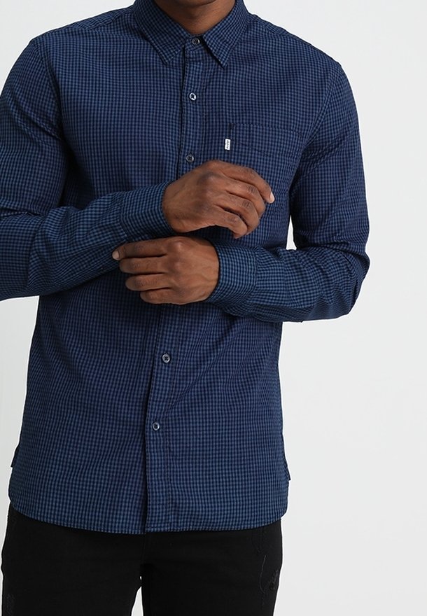 Homme portant une chemise à carreaux bleue à manches longues boutonnée, ajustant une manchette avec l'autre main sur fond blanc.